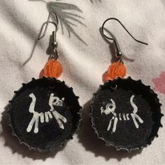 two bottle caps with cats painted on them are hanging from earrings that have orange beads