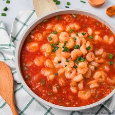 a bowl filled with shrimp and tomato soup on top of a white table cloth next to a wooden spoon