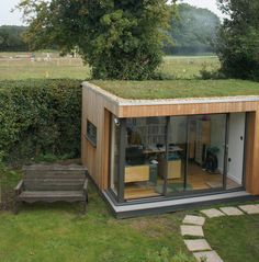 a small house with a green roof and wooden benches in the grass near some bushes