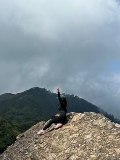 a person sitting on top of a rock with their arms up in the air and mountains behind them