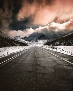 an empty road in the middle of nowhere with mountains in the background and dark clouds