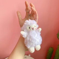 a hand holding a white stuffed animal keychain in front of a pink wall