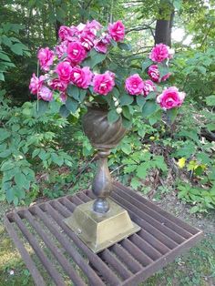 a metal vase with pink flowers in it sitting on a bench next to some trees