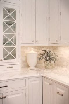 a white kitchen with marble counter tops and cabinets in the corner, vase on top