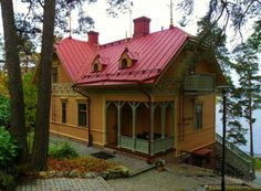 a yellow house with a red roof sitting next to trees and water in the background