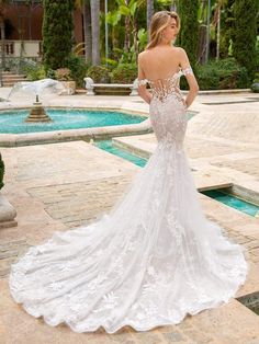 a woman in a white wedding dress standing near a pool and looking at the back of her gown