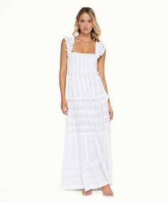 A blonde woman wearing a long white dress standing in front of a white wall. Feminine Maxi Dress With Ruffled Straps For Beach, Elegant Maxi Dress With Fitted Bodice And Ruffled Straps, Elegant Smocked Dress With Ruffled Straps, Elegant Maxi Dress With Smocked Back For Daywear, Elegant Daywear Maxi Dress With Smocked Back, Dresses With Smocked Bodice And Ruffled Straps, Elegant Beach Dress With Ruffled Straps, Elegant Beach Dresses With Ruffled Straps, Beach Maxi Dress With Square Neck And Ruffle Hem