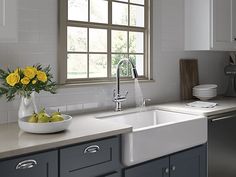 a white sink sitting under a window next to a dishwasher and a bowl of fruit