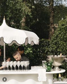 two people under an umbrella at a bar with wine glasses on the table and one person sitting behind it