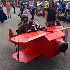 a group of people standing around a red plane with two children sitting on the wing