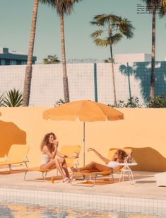 two women lounging on lounge chairs next to a swimming pool with palm trees in the background