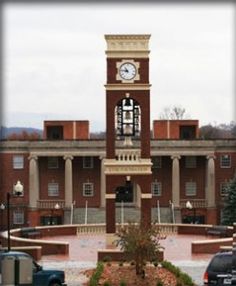 a clock tower in front of a building with cars parked around it and trees on the sidewalk