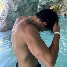 a shirtless man in the water with his hand on his head while standing next to a rock formation