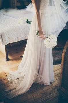 a woman standing in front of a bed wearing a wedding dress and holding a bouquet