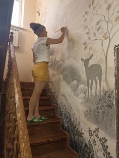 a woman painting a mural on the side of a wall with deer and trees painted on it