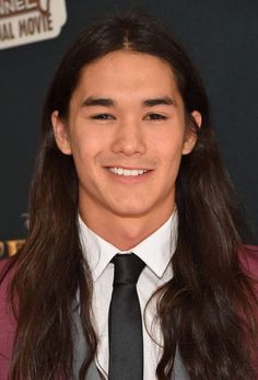 a young man with long hair wearing a suit and tie smiles at the camera while standing in front of a black background