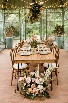 a long wooden table with flowers and greenery on it is surrounded by hanging lights