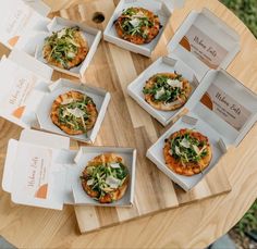 six small pizzas are sitting in boxes on a wooden table with white envelopes