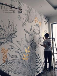 a woman painting a wall with flowers and leaves on the side, in front of a window