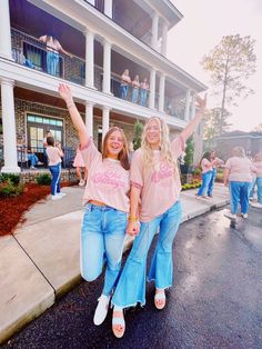 two women in pink shirts and blue jeans are walking down the street with their arms up