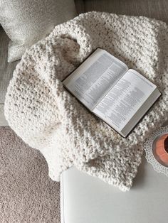 an open book on top of a white blanket next to a candle and some pillows