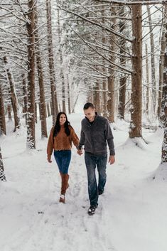 a man and woman are walking through the snow in front of some trees holding hands