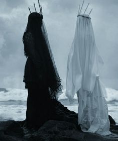 two women dressed in black and white standing on rocks near the ocean with veils over their heads