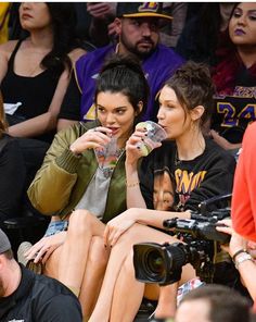 two women sitting next to each other at a basketball game, one drinking from a bottle