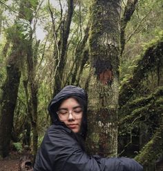 a woman in black jacket standing next to a tree with moss growing on it's trunk