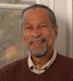 an older man wearing a sweater and tie standing in front of a window looking at the camera