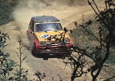 a rally car driving down a dirt road in the woods with dust coming from its tires