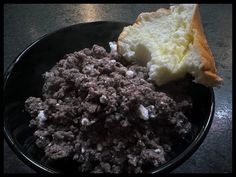 a black bowl filled with meat and bread on top of a wooden table next to a slice of bread