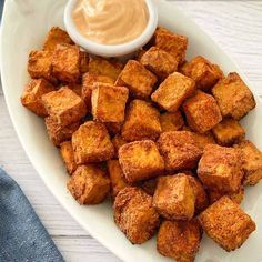 a white plate topped with tofu next to a bowl of sauce