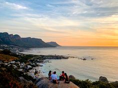 three people sitting on top of a rock overlooking the ocean