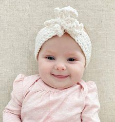 a smiling baby girl wearing a white knitted headband with a bow at the top