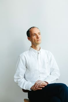 a man sitting on top of a wooden chair in front of a white wall with his hands crossed