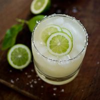 a drink with limes and sugar on the rim sitting on a wooden table next to some leaves