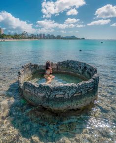 a woman is sitting in the water near an old stone bathtub at the beach