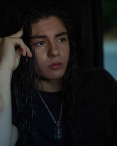 a man with long hair sitting in a car looking at the camera while holding his hand to his head