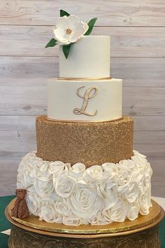 a three tiered cake with white and gold flowers on the bottom, topped with a monogram