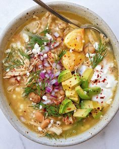 a white bowl filled with soup and topped with potatoes, onions, avocado slices and sour cream