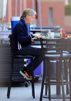 an older woman sitting at a table with food in her hand and drinking coffee on the other side