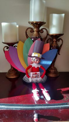 an elf is sitting on top of a table next to two candles and a paper turkey