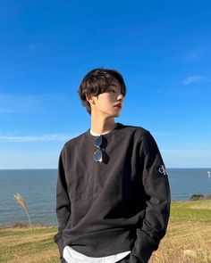 a young man standing on top of a grass covered field next to the ocean with his hands in his pockets