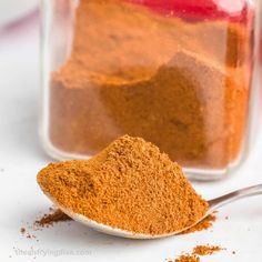 a spoon full of red powder next to a jar filled with orange colored powder on a white surface