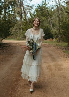 a woman walking down a dirt road holding a bouquet of flowers in one hand and smiling at the camera