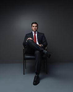 a man in a suit and tie sitting on a chair with his legs crossed, looking at the camera