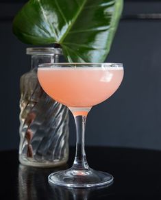 a pink cocktail in a coupe glass with a green leaf on the top and a bottle next to it