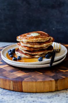 a stack of pancakes with blueberries and syrup on a wooden cutting board next to a fork