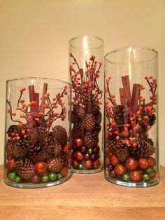 three glass vases with pine cones and berries in them sitting on a wooden table